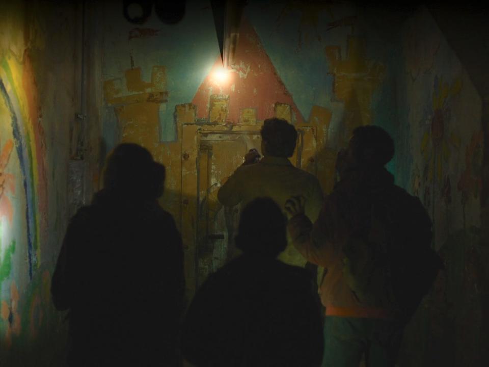 joel, ellie, henry, and sam approach a door to an underground bunker space in the last of us. the door and surrounding walls are decorated, showing a castle and a rainbow among other child-made drawings