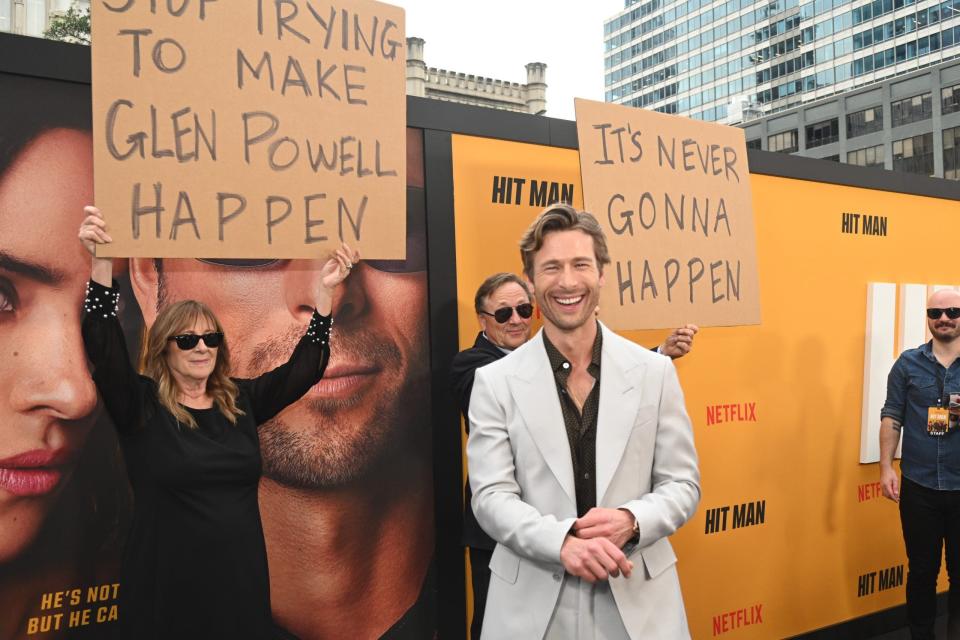 glen powell on red carpet smiling in front of parents holding signs