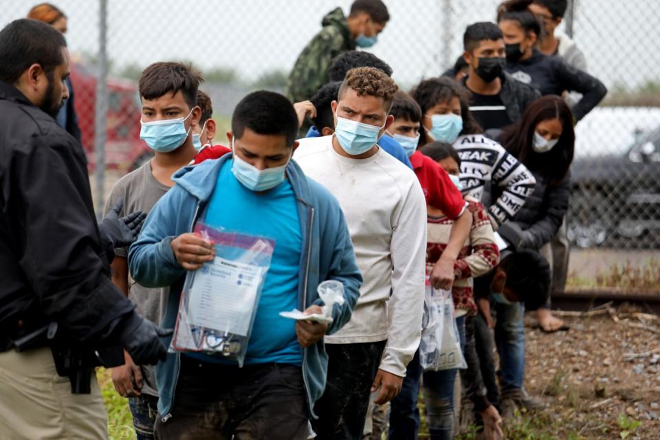 Masked people wait in line, some carrying items in clear plastic bags