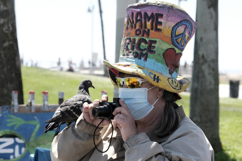 La vendedora Vivianne Robinson, que usa una mascarilla para protegerse del coronavirus, toma una fotografía mientras una paloma permanece en su brazo, cerca de la playa Venice, en Los Ángeles, el viernes 3 de julio de 2020. (AP Foto/Richard Vogel)