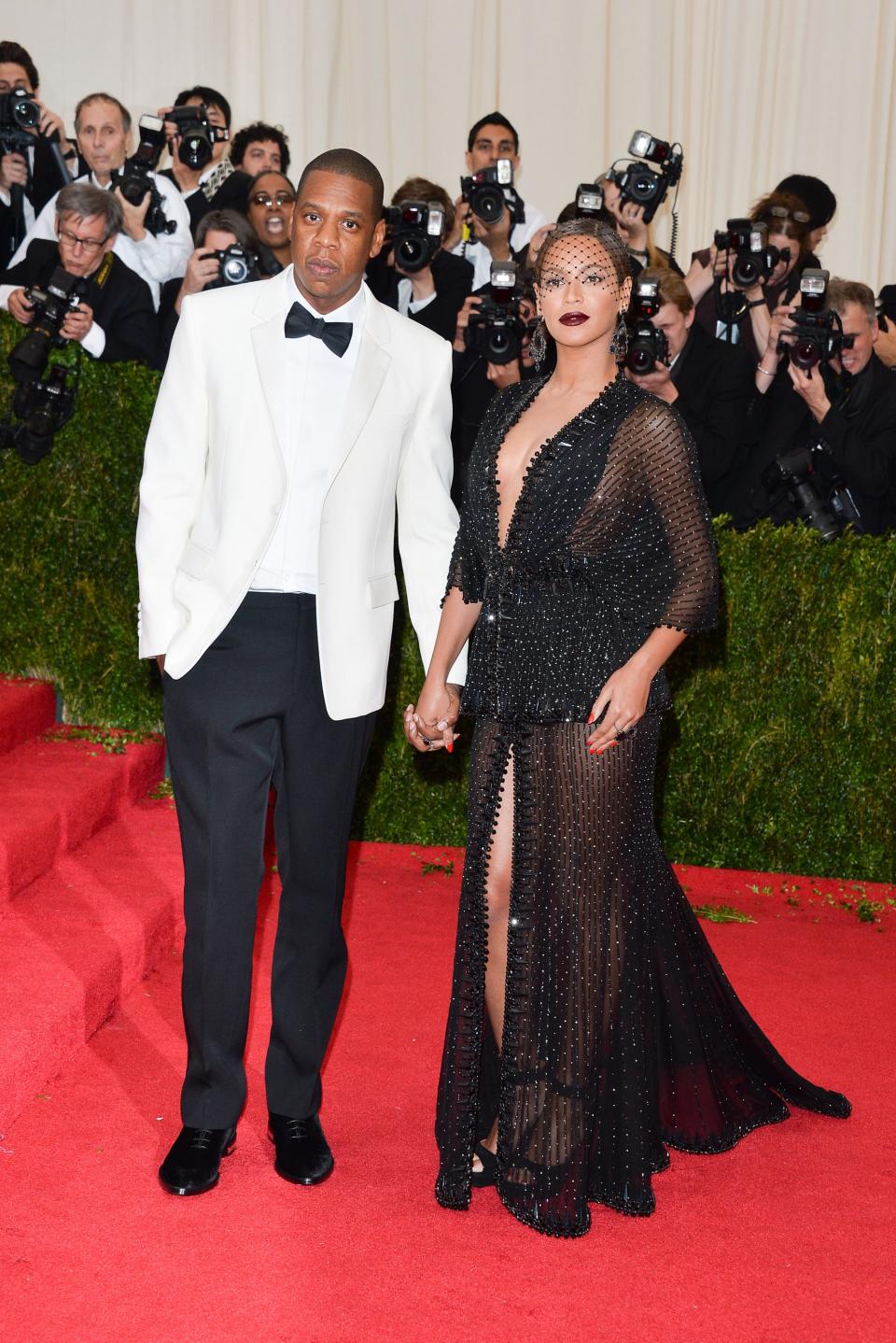 Jay-Z and Beyoncé on the red carpet at the 2014 Met Gala. Jay-Z wears a white tuxedo with black bowtie and pants. Beyonce wears a sparkling black dress with a plunging neckline and a birdcage veil.