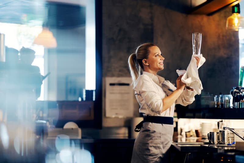 A smiling female bartender is wiping a glass while standing in a bar.