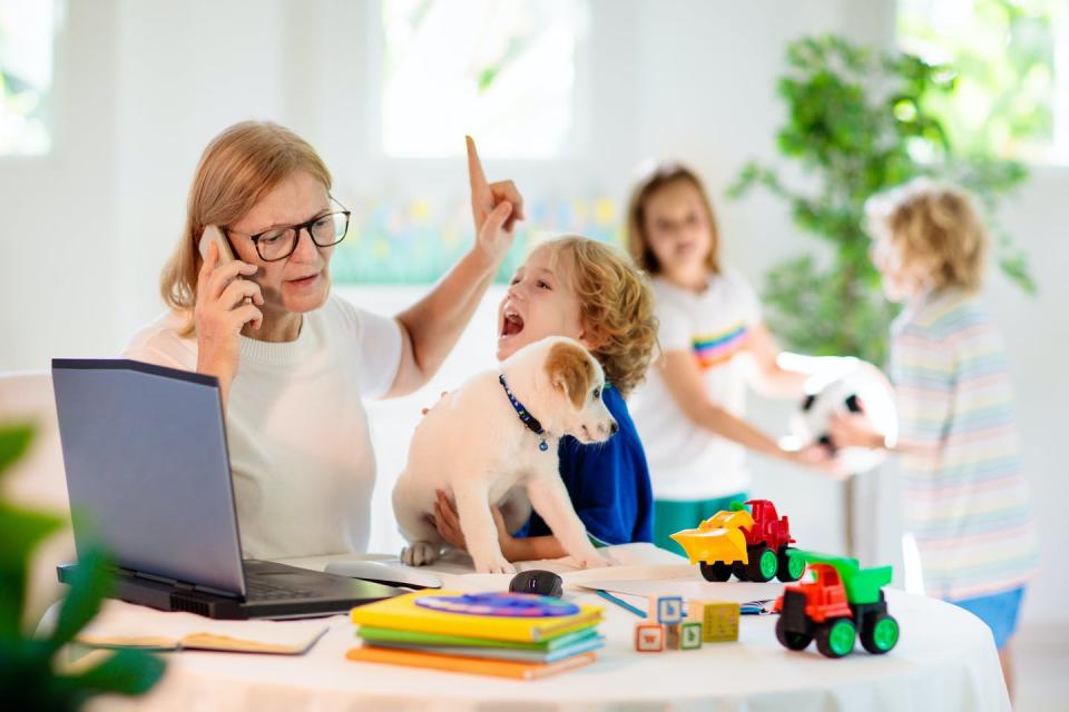 A white woman sits at a table in front of a laptop while she's on the phone and a child holds a puppy to her while two others fight over a toy in the background.