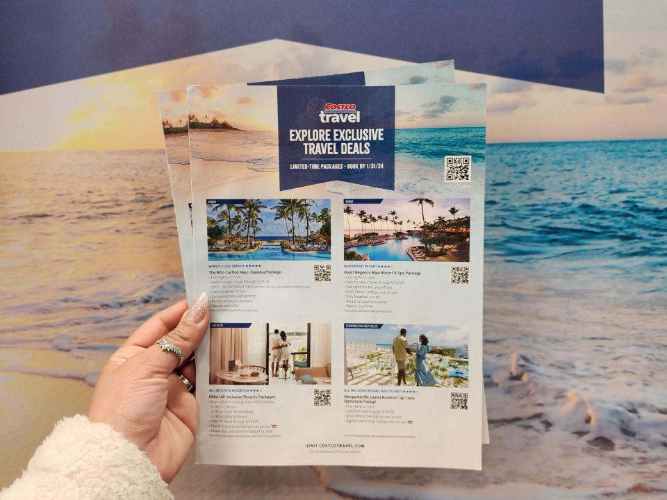 The writer holds up flyers for Costco travel deals with pictures of beaches and hotels in front of a large beach-scene backdrop