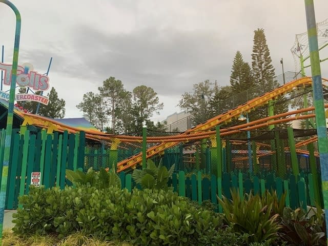 A roller coaster with multiple twists and turns is seen at an amusement park. The surrounding area has lush greenery and a wooden fence