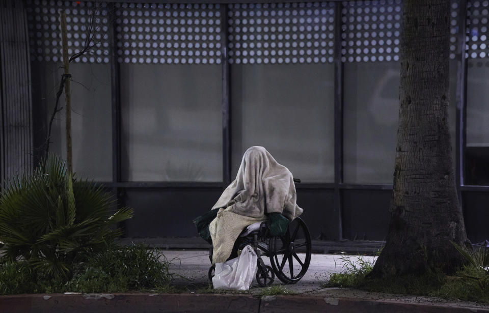 FILE - In this April 6, 2020, file photo, a homeless person sits on a wheelchair under rainy weather on Sunset Blvd., in the Echo Park neighborhood of Los Angeles. California has spent $13 billion in the last three years to tackle a massive homelessness problem likely to worsen with the pandemic, yet its approach is so disjointed and incomplete as to hinder efforts at getting people into stable housing, the state auditor said in a report released Thursday, Feb. 11, 2021. (AP Photo/Damian Dovarganes, File)