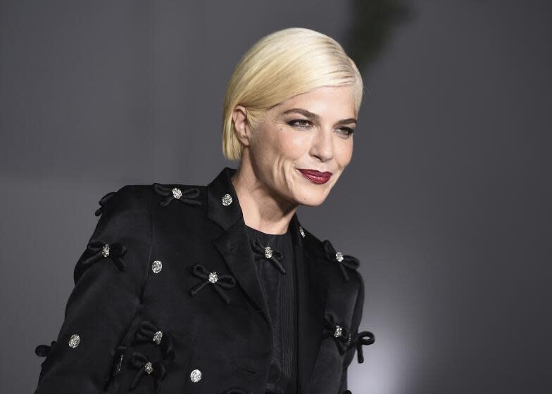 Selma Blair with short platinum hair posing in a black suit and red lipstick