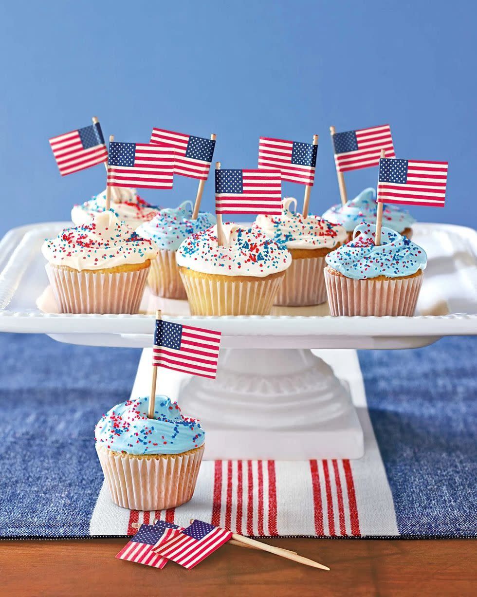 patriotic cupcakes decorated with red, white, and blue sugar and little american flags on wooden picks, all on a raised porcelain tray