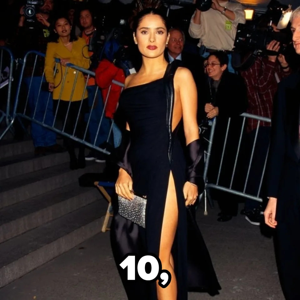 Salma Hayek poses on a red carpet in an elegant one-shoulder dress with a high slit, holding a clutch bag, surrounded by paparazzi and an audience