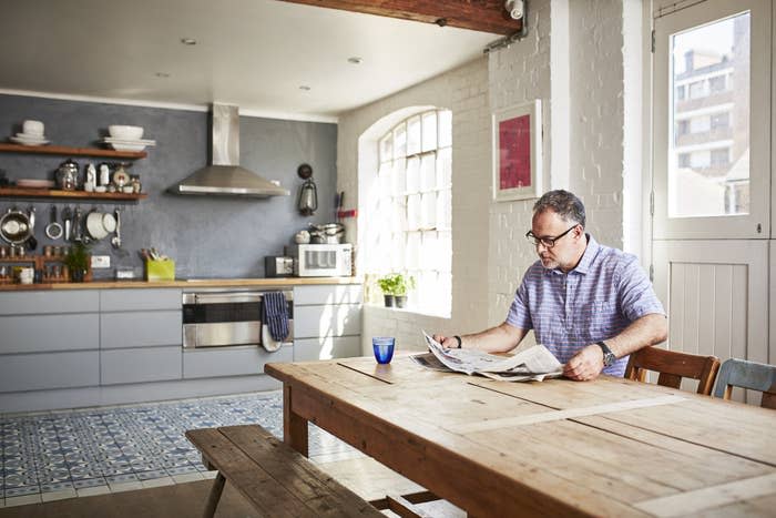 A middle-aged man sits at a wooden table and bench in a large kitchen, reading a newspaper