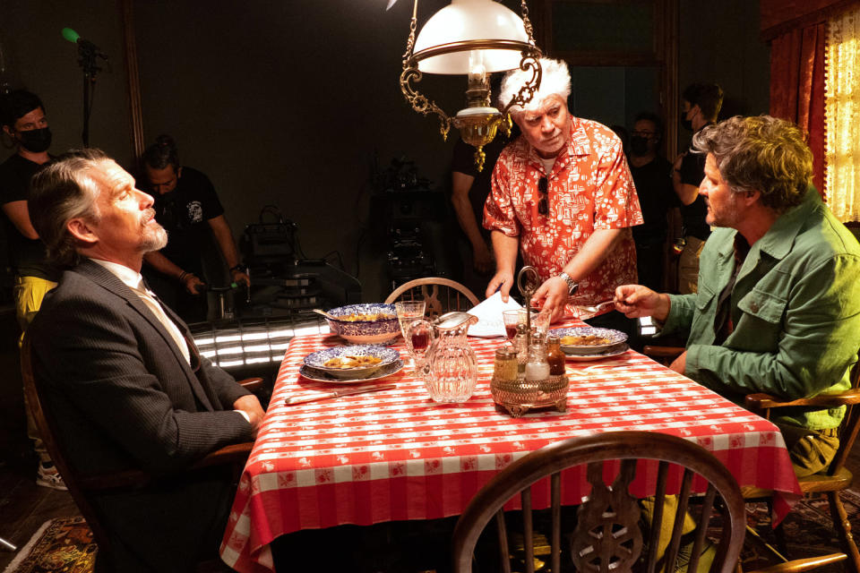 Pedro Almodóvar works with Ethan Hawke and Pedro Pascal at a dinner table while crew members hold gear in the background on the set of 