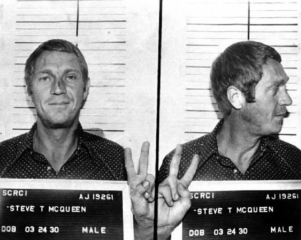 Mugshot of Steve McQueen with front and side profiles, holding a name placard; he gestures a peace sign with his hand