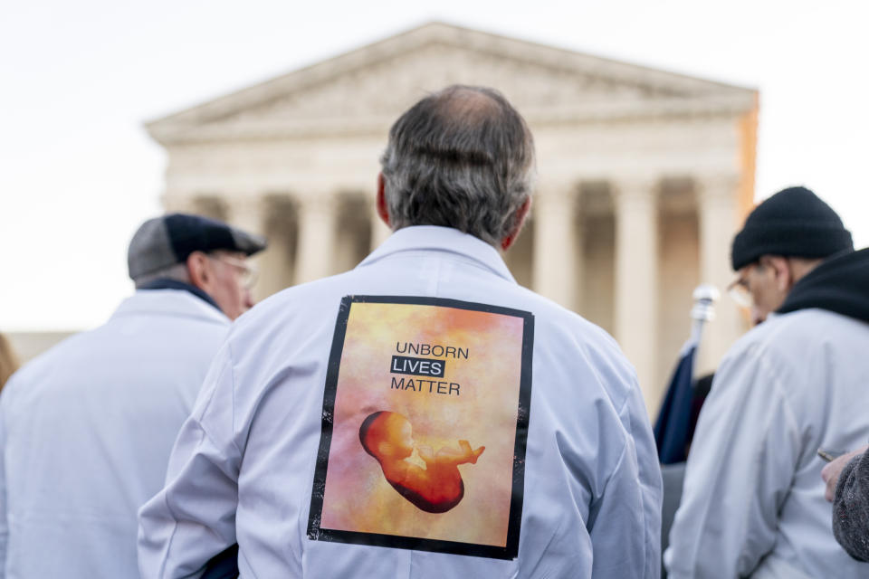 A man wears a sign on his back that reads "Unborn Lives Matter" as he and other anti-abortion protesters wearing doctors uniforms demonstrate in front of the U.S. Supreme Court, Wednesday, Dec. 1, 2021, in Washington, as the court hears arguments in a case from Mississippi, where a 2018 law would ban abortions after 15 weeks of pregnancy, well before viability. (AP Photo/Andrew Harnik)