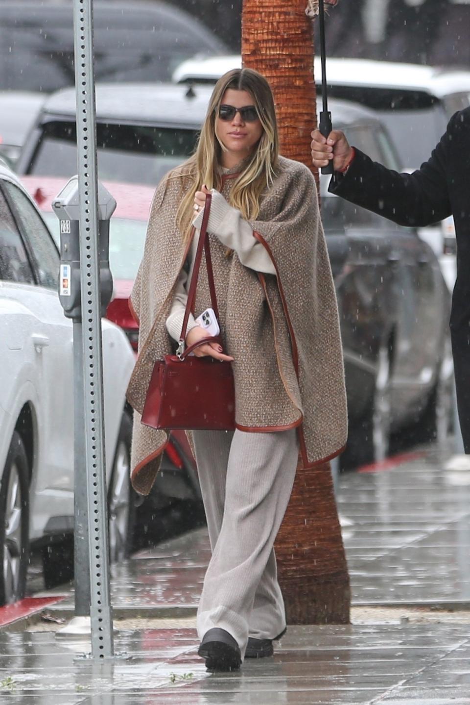 Sofia Richie Grainge wearing a tan poncho with loose fitting pants and a burgundy handbag