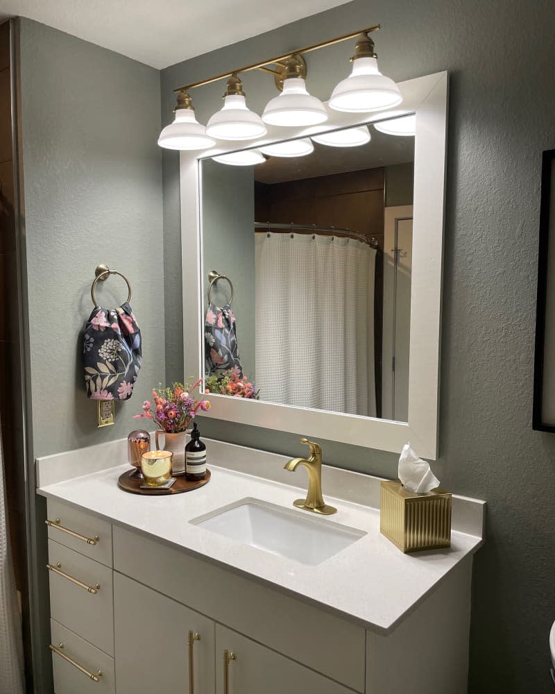 Wood tray holding accessories and gold tissue box atop white sink with gold faucet and white cabinetry under white framed mirror in sage bathroom with floral towel.