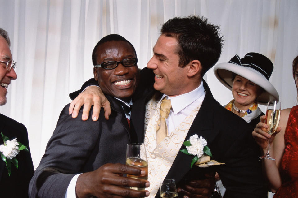 Two men, dressed in suits with boutonnières, smile and embrace while holding champagne glasses at wedding