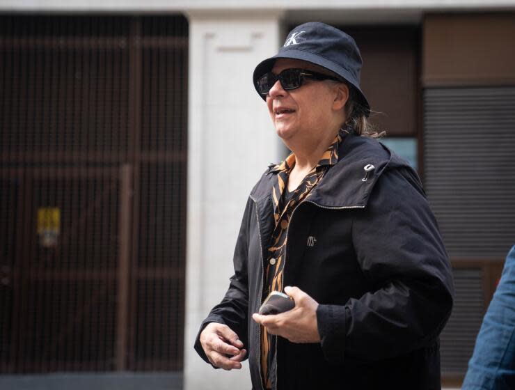 Duran Duran's Andy Taylor walks and smiles while wearing a black bucket hat and jacket and a brown and black shirt