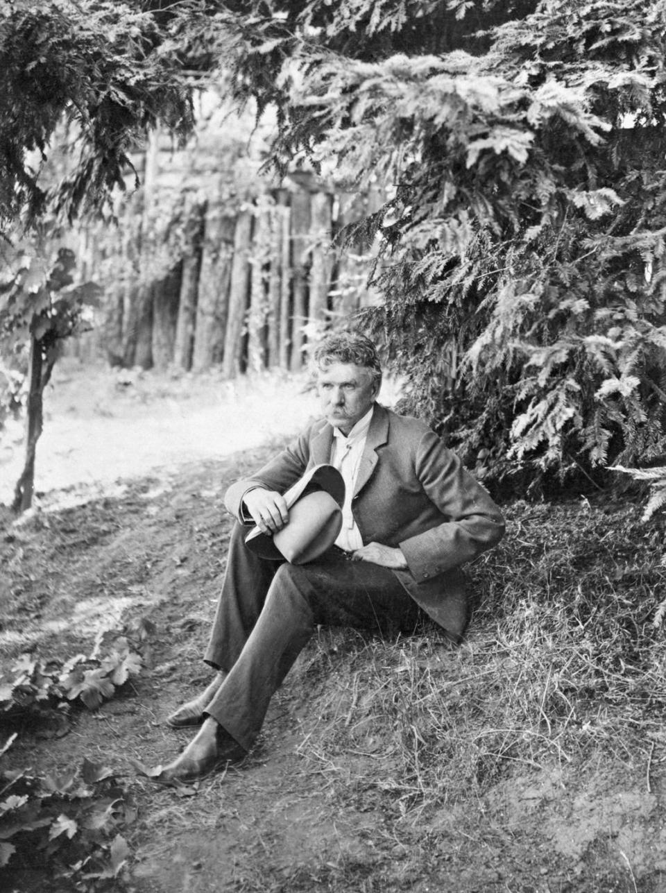 ambrose bierce seated under tree holds his hat in one hand, he wears a suit