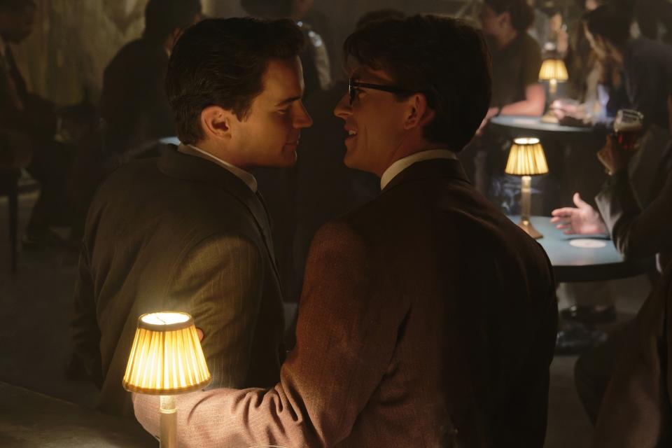 Matt Bomer and Jonathan Bailey, dressed in suits, share an intimate moment at a dimly-lit table in a crowded venue