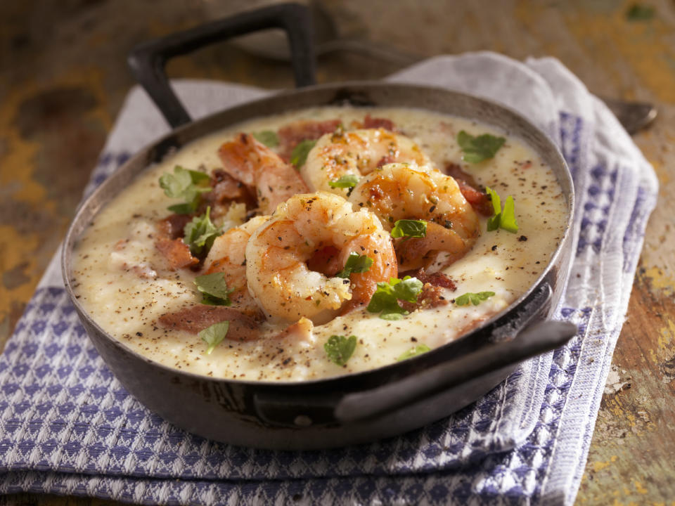 A cast iron skillet filled with grits topped with shrimp, bacon pieces, and green garnish, on a folded gingham-style cloth napkin on a wooden table