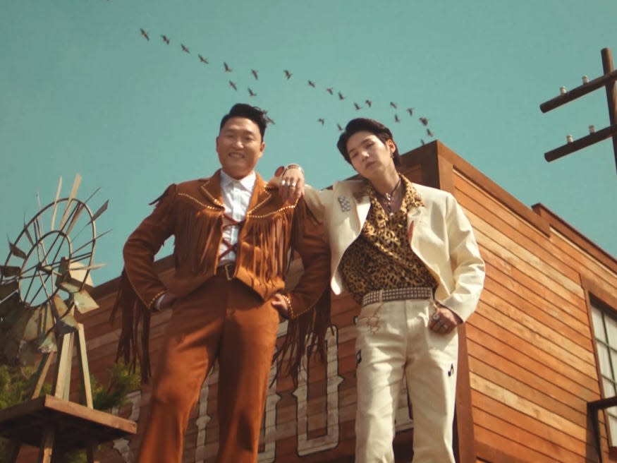 two men standing together on top of a car. the one on the left (psy), slightly older, is wearing a brown suit with fringe on the front. the one on the left (suga), slightly younger, is wearing a white suit with a cheetah print shirt, his arm resting on the other man's shoulder