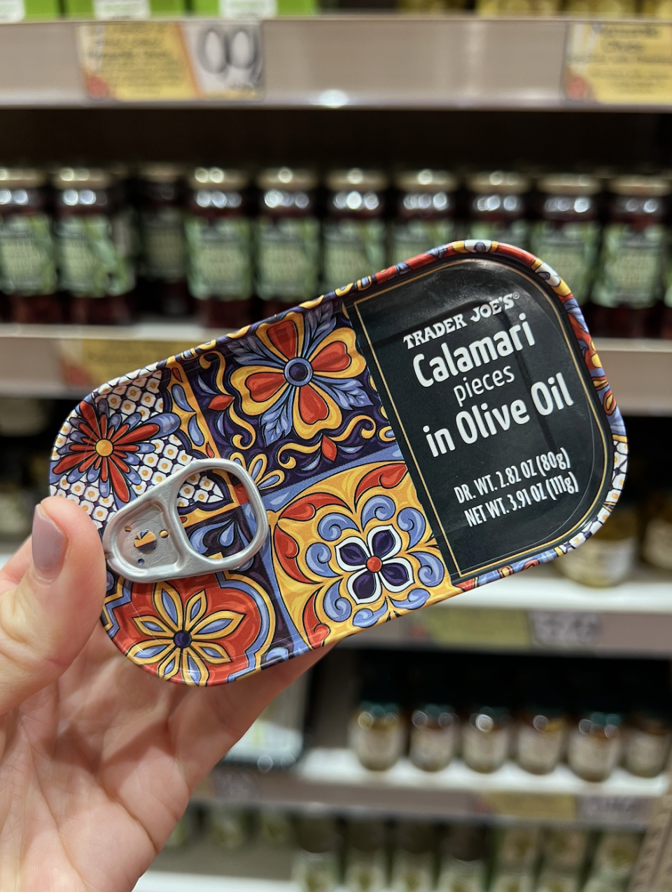 Hand holding a can of Trader Joe's Calamari pieces in Olive Oil, with a colorful floral design, in front of a grocery store shelf filled with various canned goods