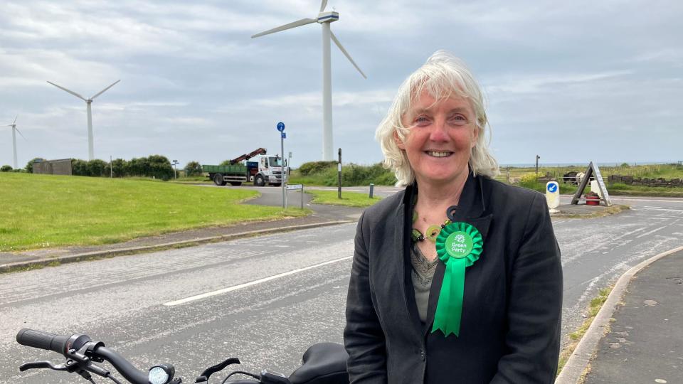 Woman with long grey hair wearing green badge and standing outside wind turbine