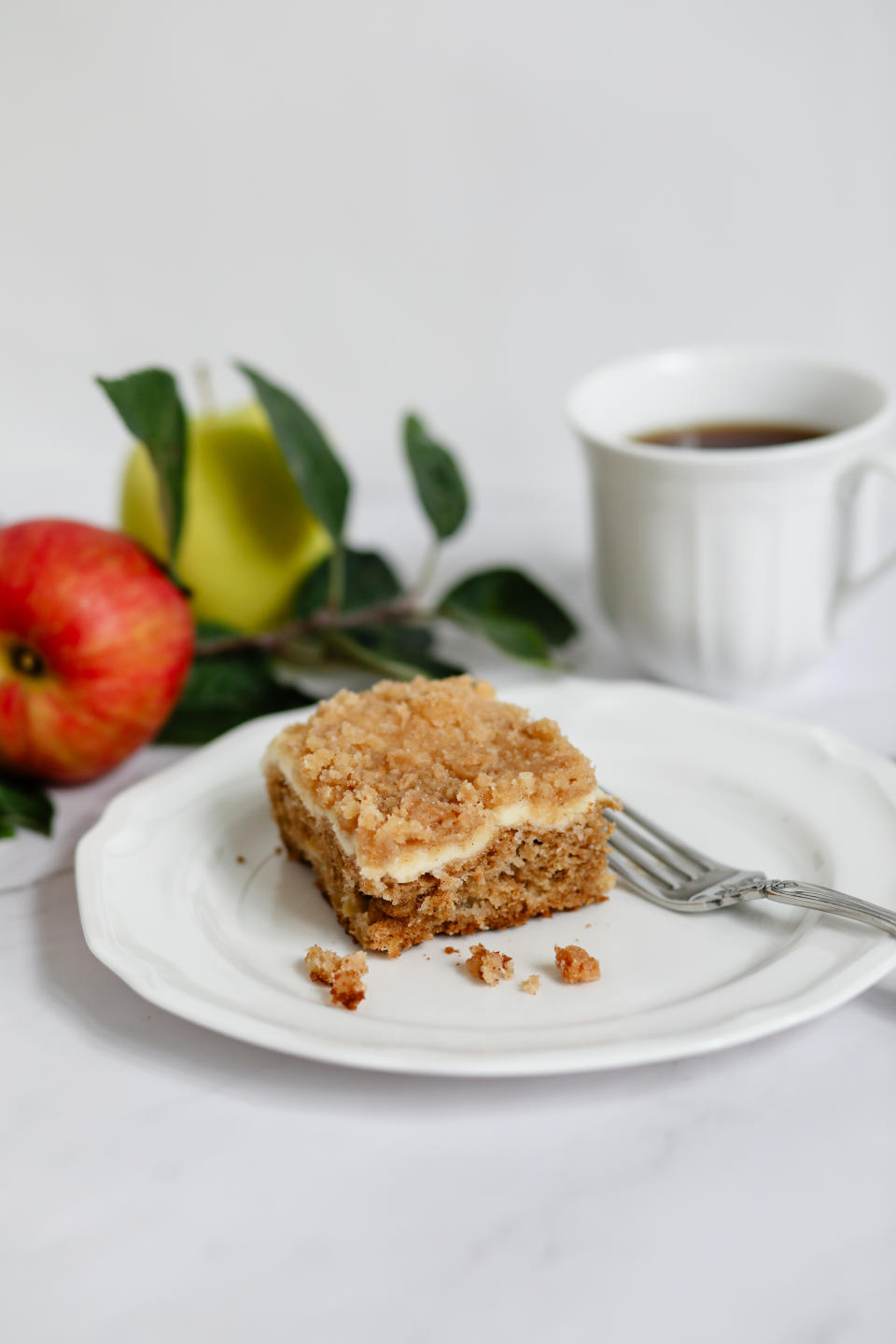 Slice of crumb cake on a white plate with a fork, accompanied by an apple, a green fruit, and a cup of coffee