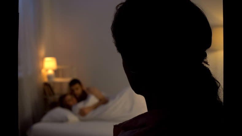 Silhouette of a person in the foreground with a couple embracing in a bed in the background