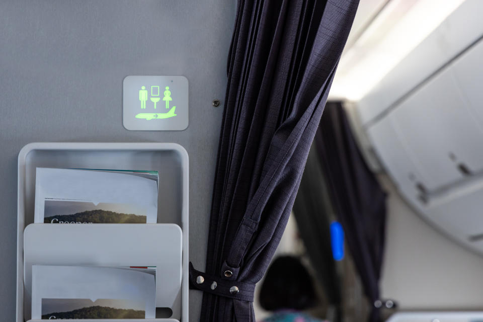 Interior of an airplane showing seat with curtain, overhead restroom sign, and seatback pocket with brochures