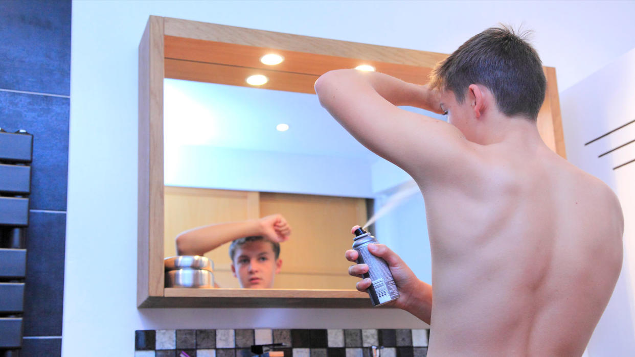  Young teen boy using a spray deoderant on his left armpit while he looks in a bathroom mirror. 