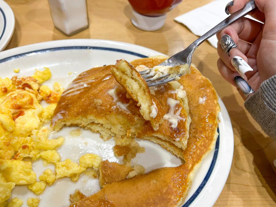 Stack of two pancakes cut in half and a fork with a piece of pancakes above it. Scrambled eggs with ketchup sit on the plate