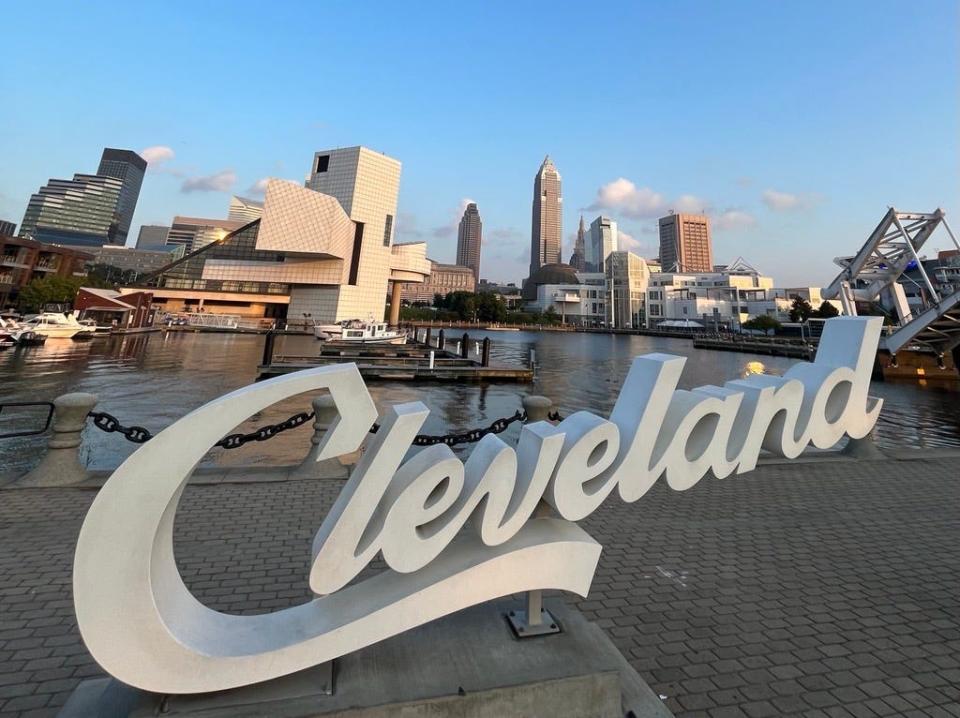 A Cleveland script sign sits on the North Coast Harbor with the Rock & Roll Hall of Fame and the Cleveland skyline in the background.