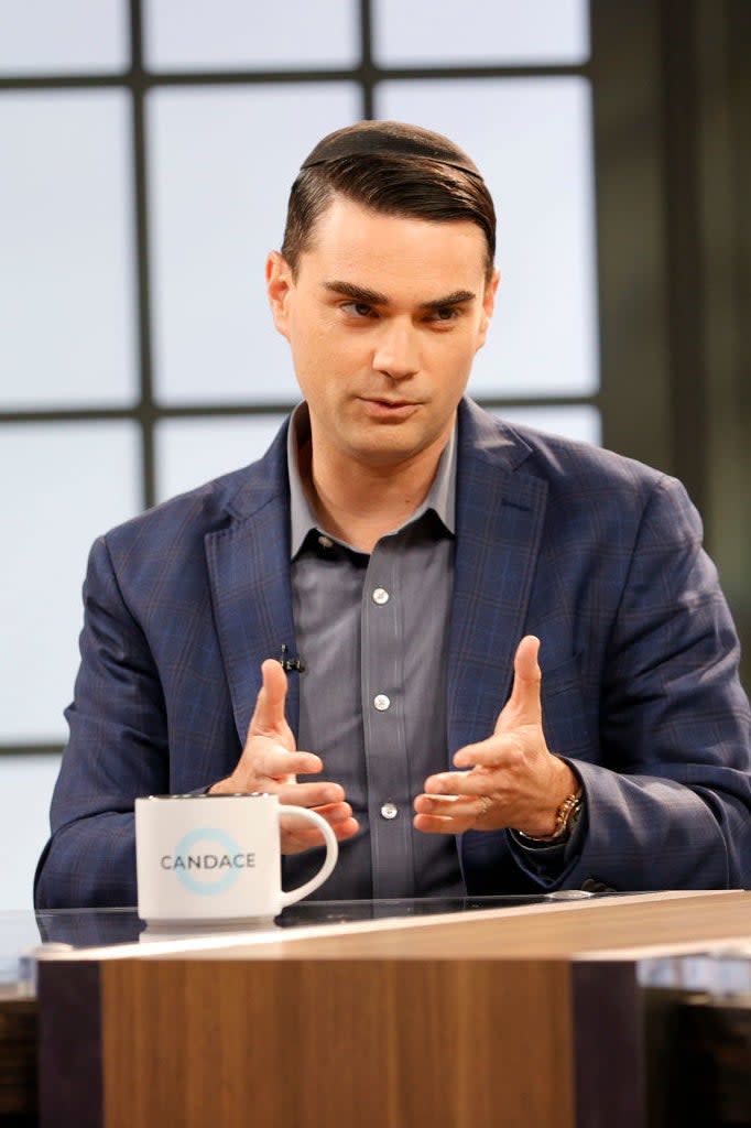 Man in a plaid suit speaking with hands open on a talk show set with a mug labeled 'CANDACE'