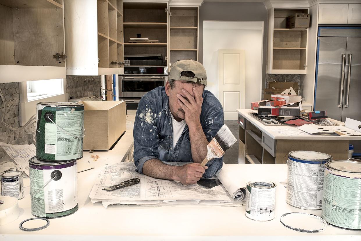 Male painter, face in hands, wearing clothes covered in paint and hold a wet paint brush in messy home kitchen during remodel project with empty paint cans stacked everywhere on kitchen counter with lids off