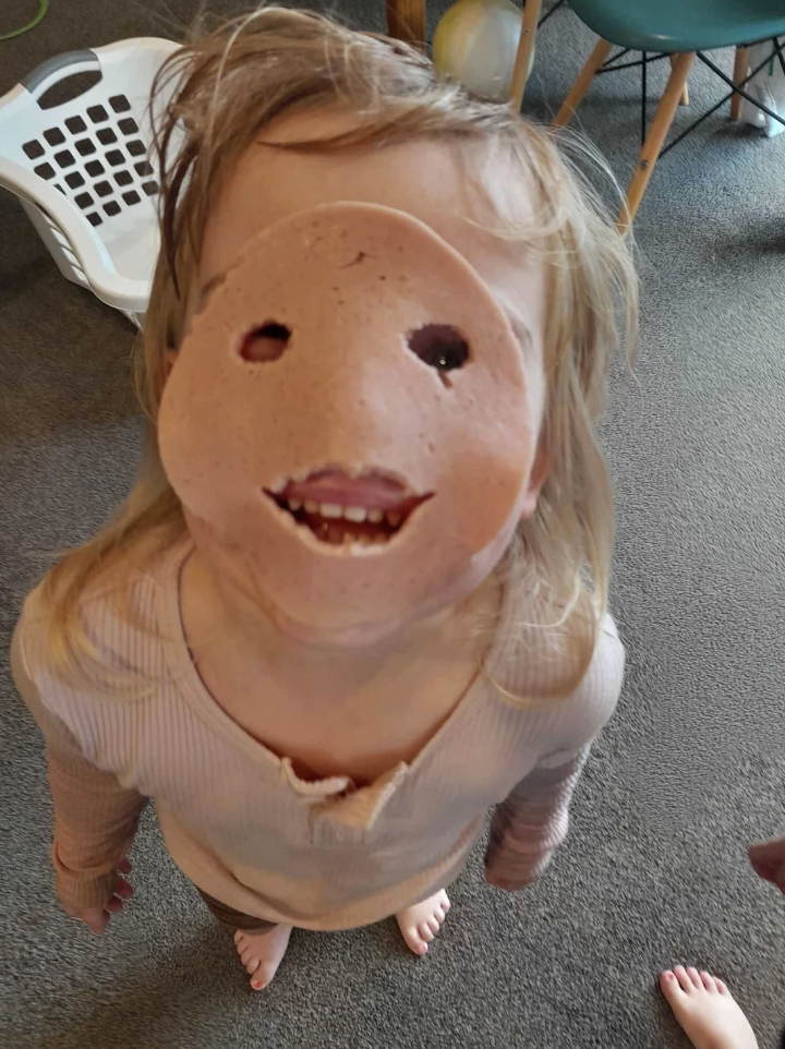 Child wearing a large mask of a happy face, standing indoors with furniture around