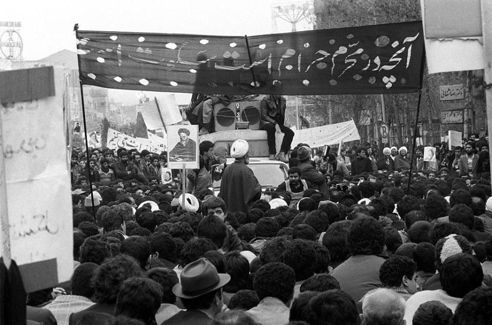 Black-and-white photo of a large crowd around a military vehicle, posters showing the ayatollah, and signs in Persian