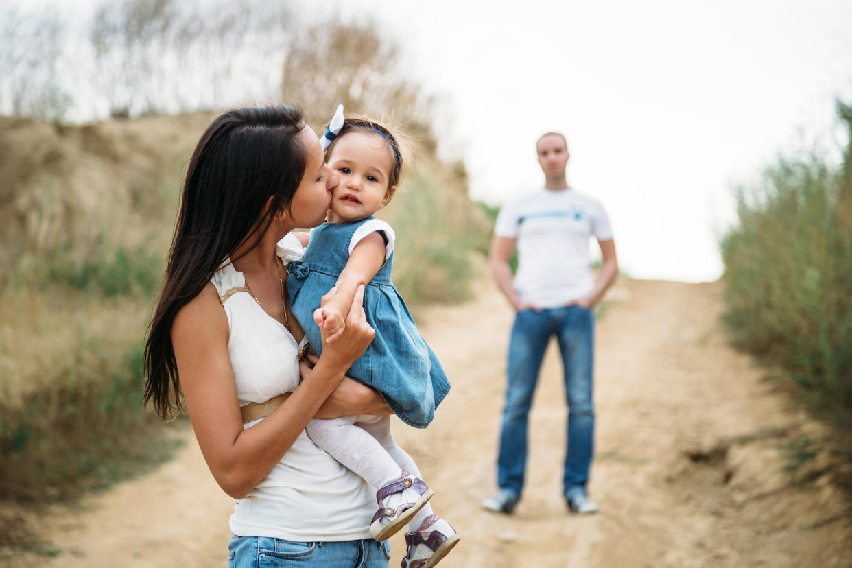 Young mother in white t-shirt and blue jeans with a small daughter in dress hugging and kissing with a father on the background.