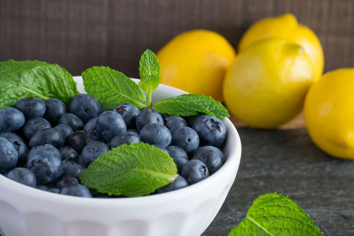 Blueberries in a white bowl with mint, lemon, almonds - healthy ingredients on a black slate board with wooden background. Bright colors for organic and health lifestyle choice.