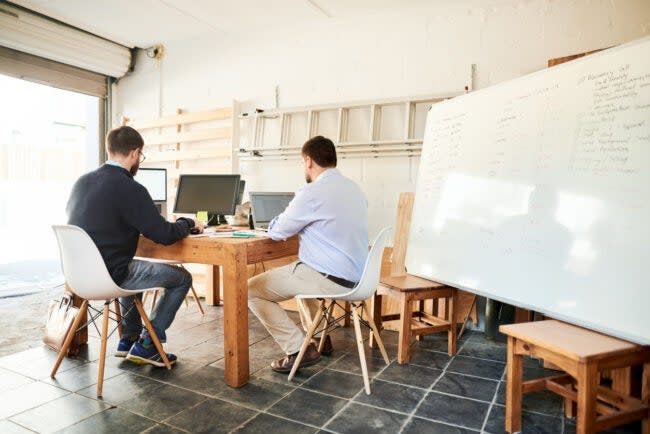 Two men working at a wooden table in home office/converted garage with a whiteboard next to the table.