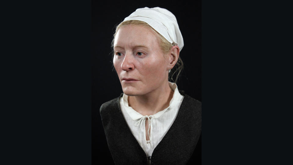 We see the bust reconstruction of a woman wearing a gray jacket, white blouse and has her head covered by a white cap.