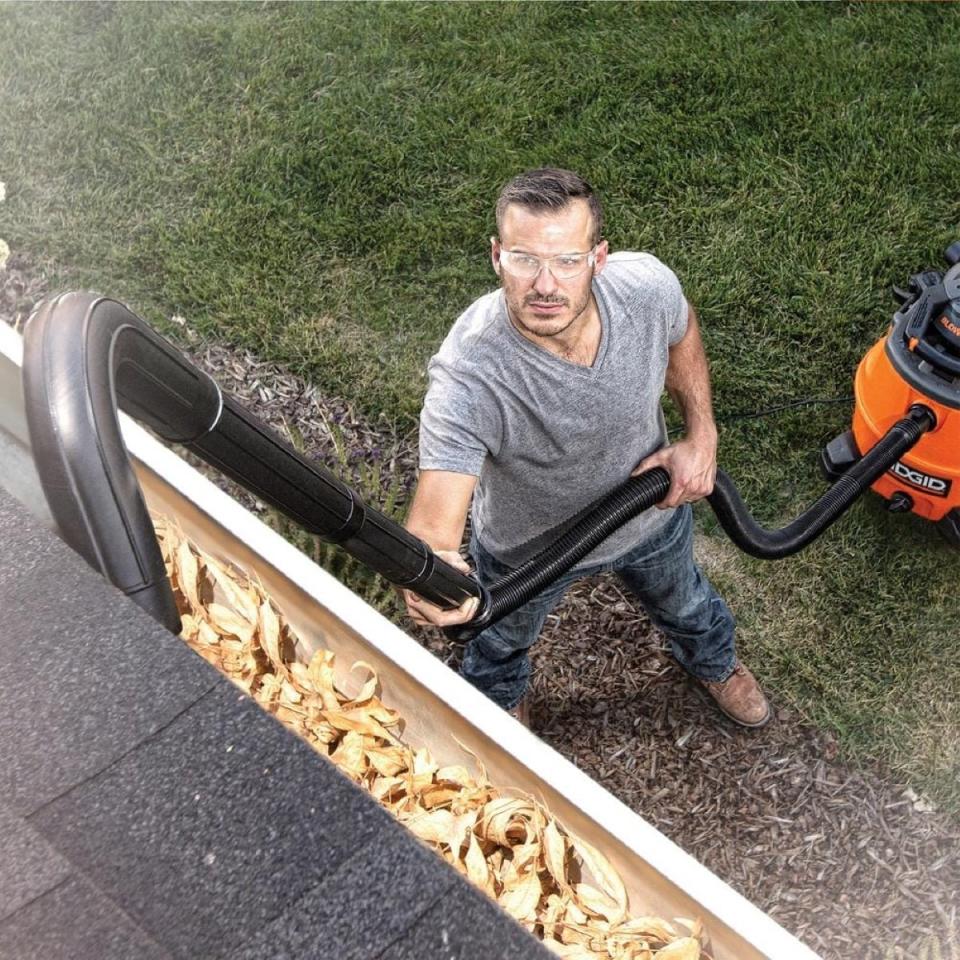 Man wearing safety glasses using a wet/dry vacuum with hose attachment to clean rain gutters.