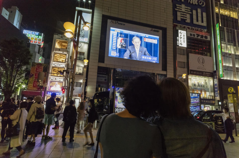 FILE -In this May 28, 2021, file photo, a couple watches a big screen as Japanese Prime Minister Yoshihide Suga speaks in Tokyo. With infections and serious cases rising despite a state of emergency, Suga has begun all-out efforts, from deploying military doctors and nurses at mass inoculation centers to allowing legal exceptions to recruit more people to give shots. But the realization here that it may be too little, too late to make a difference during the Olympics is starting to dawn. (AP Photo/Hiro Komae, File)