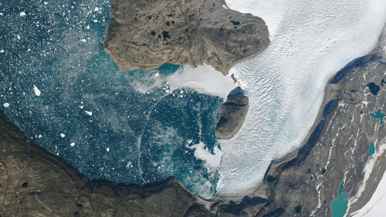  A satellite photo of a fjord with water covered in tiny icebergs and an wave of water arcing across the surface. 