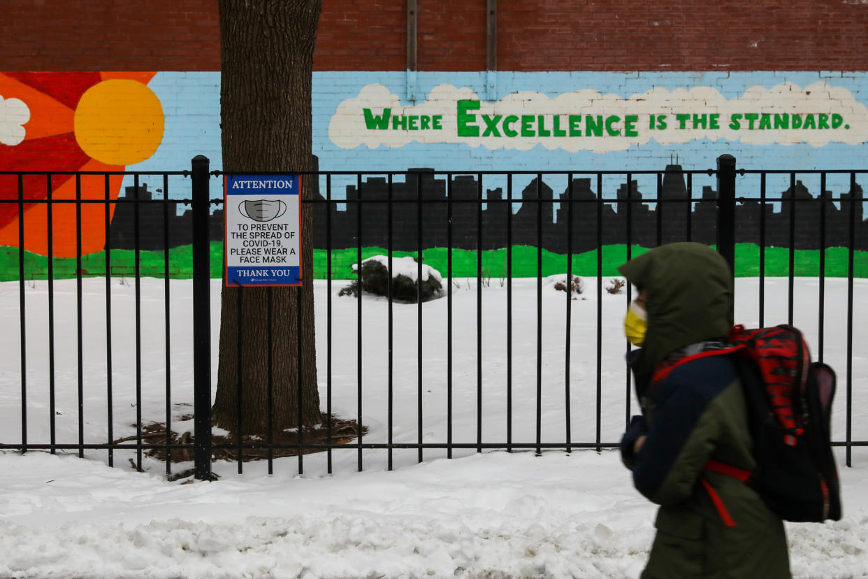 A masked student in heavy outerwear and backpack walks through snow past a sign recommending the use of face masks to prevent the spread of COVID-19. On the school wall, a sign reads: 