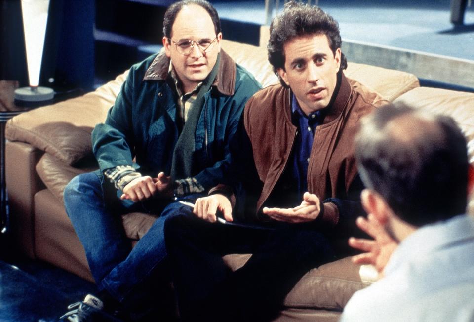 Jason Alexander and Jerry Seinfeld sit on a couch, talking to a man whose back is to the camera