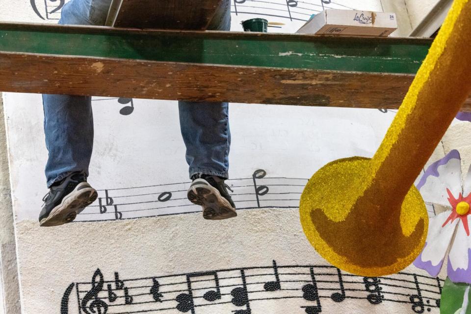 A volunteer sits on scaffolding while decorating a float that includes an image of musical notes and a horn.