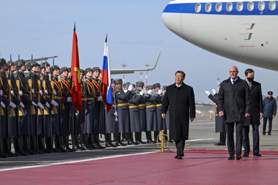 A man in a dark overcoat, center, walks past people in military uniform lined up near two flags. On the right is an airplane