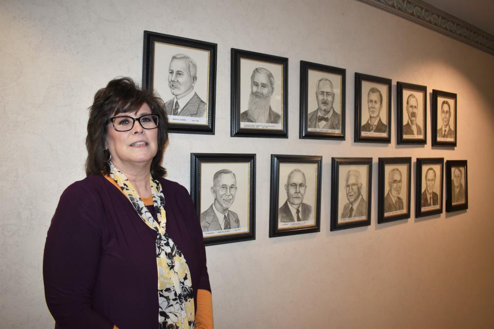 Blissfield State Bank President and CEO Julie Goll stands alongside the photos of the bank's former and past presidents Friday morning. Goll is Blissfield State Bank’s first woman president and CEO. She is in her ninth year as president and is in her 41st year with the bank.