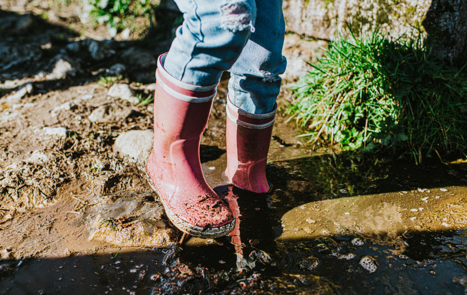 Child in bright pink rain boots wades though a deep muddy puddle. One boot sinks down into the soft mud. Space for copy.